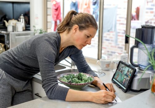 Woman in the kitchen watching zoom-meeting on tablet and writes notes